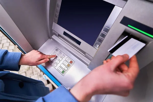 Kéz bankautomatánál gombot nyom, a másik kéz bankkártyát tart a kártyanyílás mellett.